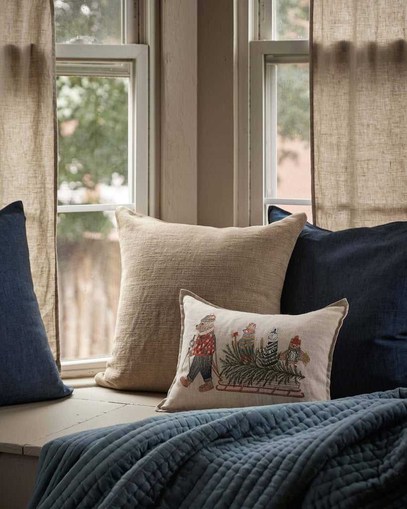 A cozy window seat with beige and navy blue pillows, a quilted blue blanket, and the Coral & Tusk Christmas Tree Sled Pillow embroidered with children sledding. Light brown curtains frame the window, overlooking trees outside.