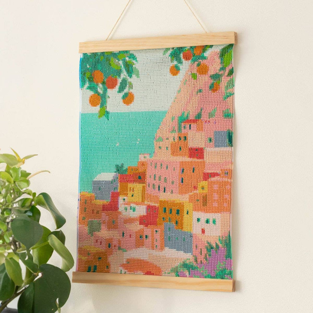 The Diamond Painting Kit - Positano by Petra Lidze features a vibrant coastal hillside town with pastel buildings, lush greenery, orange trees, a turquoise sea, and a potted plant in the foreground.