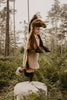 A young child in a Brown Horse costume stands on one leg atop a round, fluffy stool in a forest, surrounded by green ferns and tall trees.