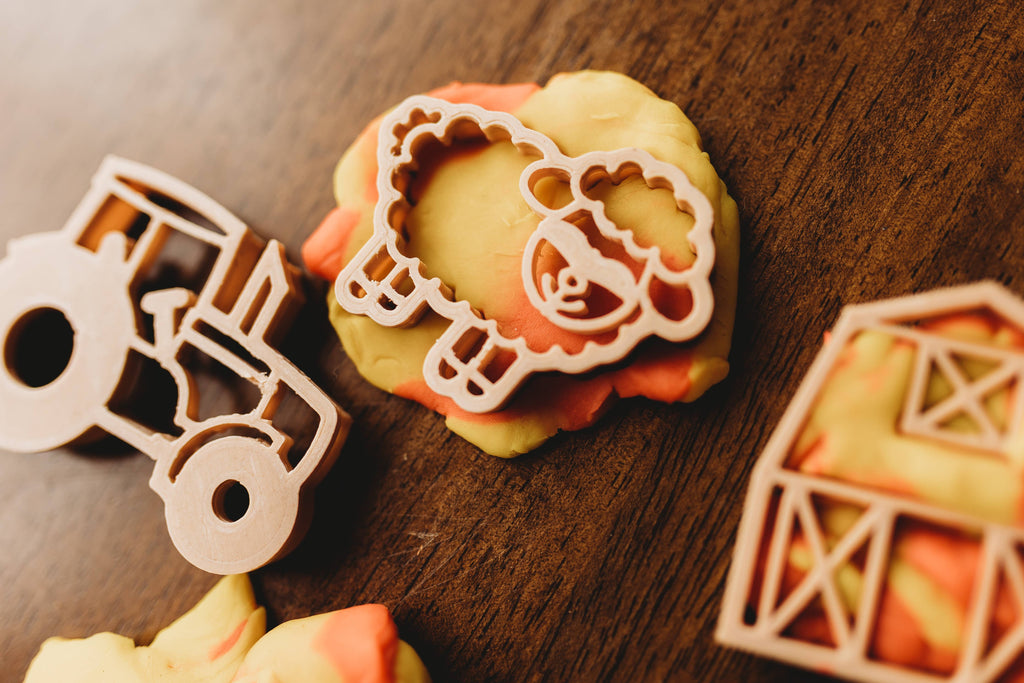 The Farm Cutter Set - Eco Playdough Tool features plastic cookie cutters shaped like a sheep, a tractor, and a barn, resting on marbled orange and yellow dough atop a wooden surface—ideal for inspiring sensory play for little hands.