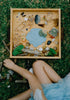 A child sits on grass by a wooden tray filled with toy animals, stones, leaves, and a blue clay pond. The playful, nature-themed scene features creations made with the Pond Life Cutter Set – Eco Playdough Tool, crafted from biodegradable PLA.