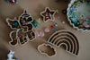 The Unicorn Cutter Set – Eco Playdough Tool, featuring plastic molds of a unicorn, star, rainbow, and cloud, is arranged on a brown surface surrounded by colorful beads and gems. This eco-friendly playdough tool set is perfect for sensory play, with a unicorn toy partly visible in the corner.