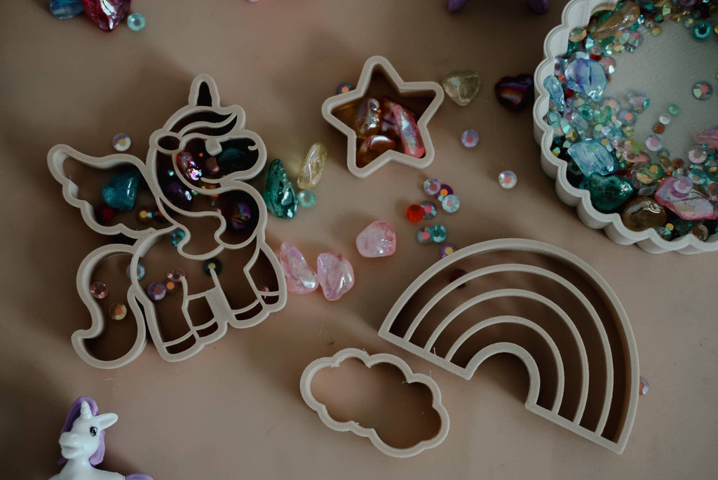 The Unicorn Cutter Set – Eco Playdough Tool, featuring plastic molds of a unicorn, star, rainbow, and cloud, is arranged on a brown surface surrounded by colorful beads and gems. This eco-friendly playdough tool set is perfect for sensory play, with a unicorn toy partly visible in the corner.