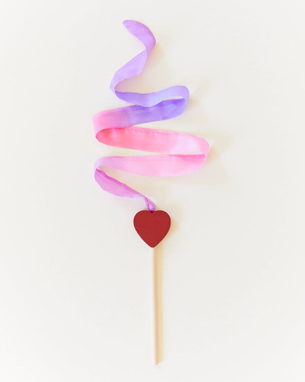 A minimalist artistic representation of a Sarah's Silk Mini Play Heart Streamer Wand, featuring rainbow streamers in pastel purple and pink shades, forming a spiral, topped with a wooden stick that has a red heart attached at its end.