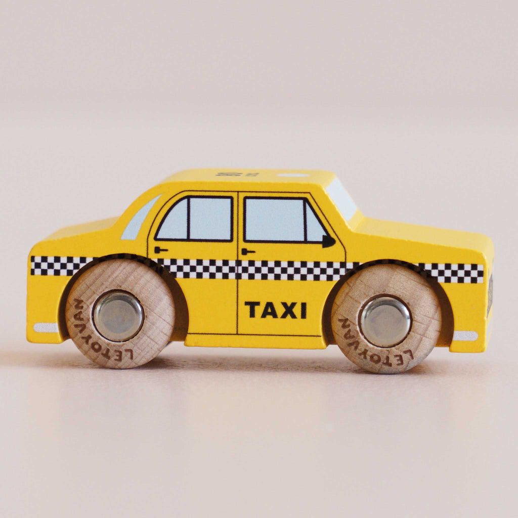 A yellow wooden No. 39 GT245 Trixie taxi toy, made in England, featuring classic black and white checkered detailing and “TAXI” on the side—shown in profile on a light background—perfect for imaginative play inspired by an iconic New York cab.