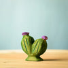 A small ceramic cactus figurine from the Bumbu Wooden Cacti Set features two green stems topped with purple blooms. It sits on a light wooden surface against a soft blue background, showcasing detailed textures and white dots.