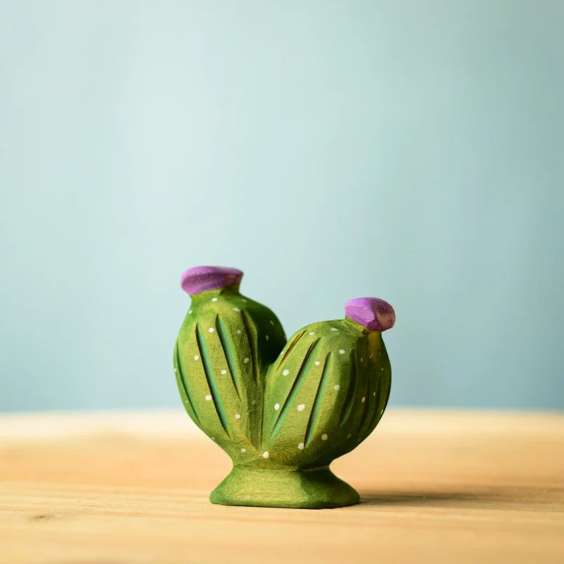 A small ceramic cactus figurine from the Bumbu Wooden Cacti Set features two green stems topped with purple blooms. It sits on a light wooden surface against a soft blue background, showcasing detailed textures and white dots.