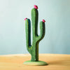 A small, green, decorative cactus with white spots and three pink buds from the Bumbu Wooden Cacti Set is placed on a light wood surface. The background is a soft, gradient blue, providing a calm and minimalist backdrop.