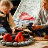 Two children are sitting on the floor playing with a Campout Campfire & S'mores. The campfire, made of faux felt logs and fabric flames, is surrounded by soft, gray stones. They're making pretend s’mores over the fire. Behind them, there's a tent featuring a world map design. The scene is cozy and playful.