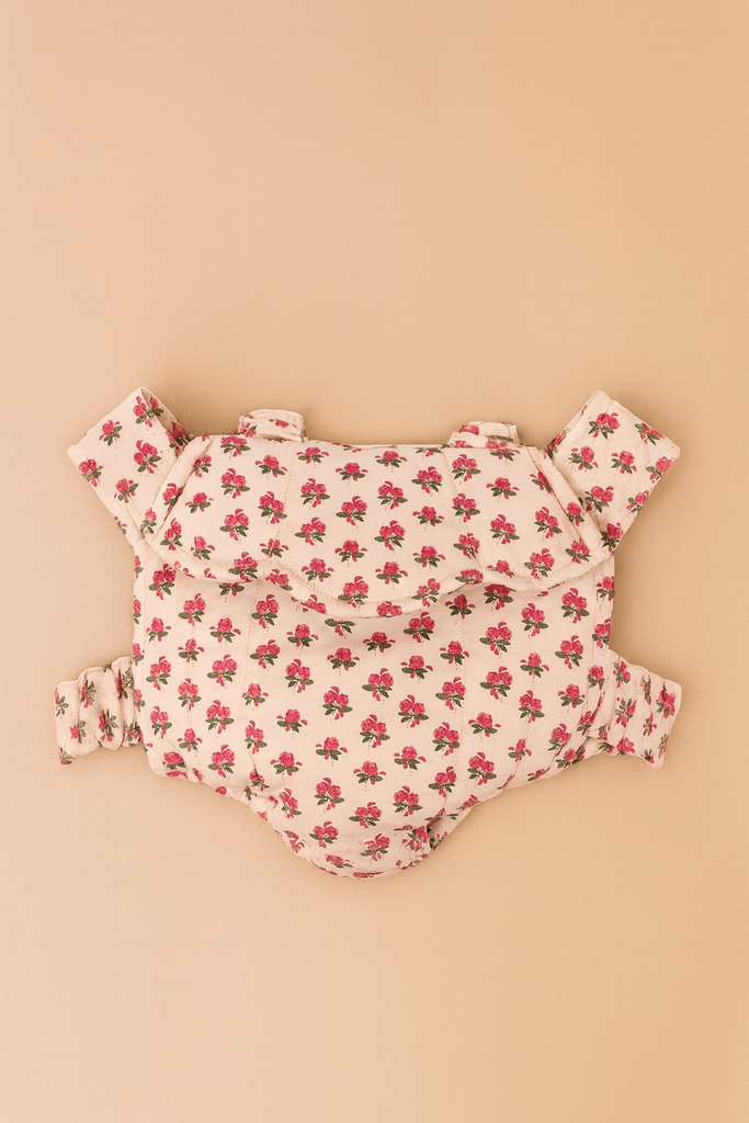 The Konges Sloejd Doll Carrier - Rosie Rose features a soft, floral cotton design with pink and green flowers on cream, padded straps, and structured support—a charming toy accessory shown flat on a beige surface.