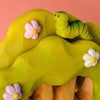 The Bumbu Handmade Wooden Caterpillar, featuring a smiling face, rests atop a painted green wooden tree adorned with pastel pink and white flowers. Set against a pink background, this charming toy invites imaginative play.