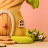 A wooden treehouse with a round door and small window stands next to a Bumbu Handmade Wooden Caterpillar toy and purple flowers, inviting imaginative play. The background features a soft pink gradient.