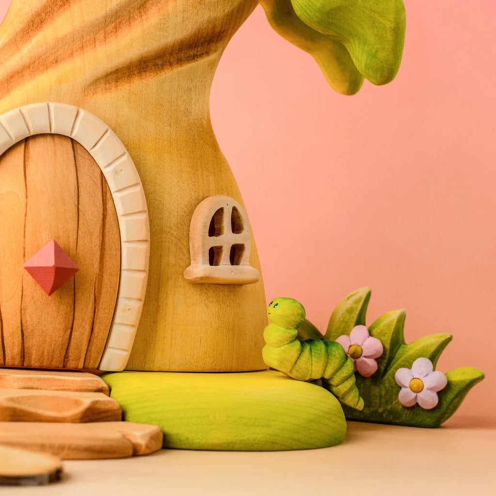 A wooden treehouse with a round door and small window stands next to a Bumbu Handmade Wooden Caterpillar toy and purple flowers, inviting imaginative play. The background features a soft pink gradient.
