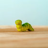 The Bumbu Handmade Wooden Caterpillar, a small green wooden toy with painted eyes, rests on a light wooden surface against a pale blue background, inviting imaginative play and serving as an appealing Montessori toy for young children.