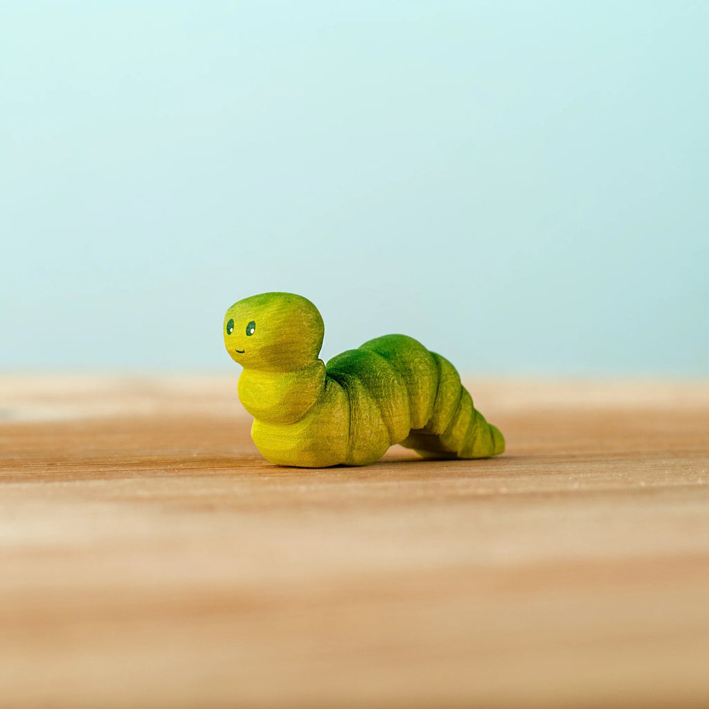 The Bumbu Handmade Wooden Caterpillar, a small green wooden toy with painted eyes, rests on a light wooden surface against a pale blue background, inviting imaginative play and serving as an appealing Montessori toy for young children.