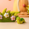 The Bumbu Handmade Wooden Caterpillar, painted in green, rests beside a decorative green piece adorned with white and pink flowers—inviting imaginative play. The softly blurred background features pastel colors and shapes, creating a whimsical scene.
