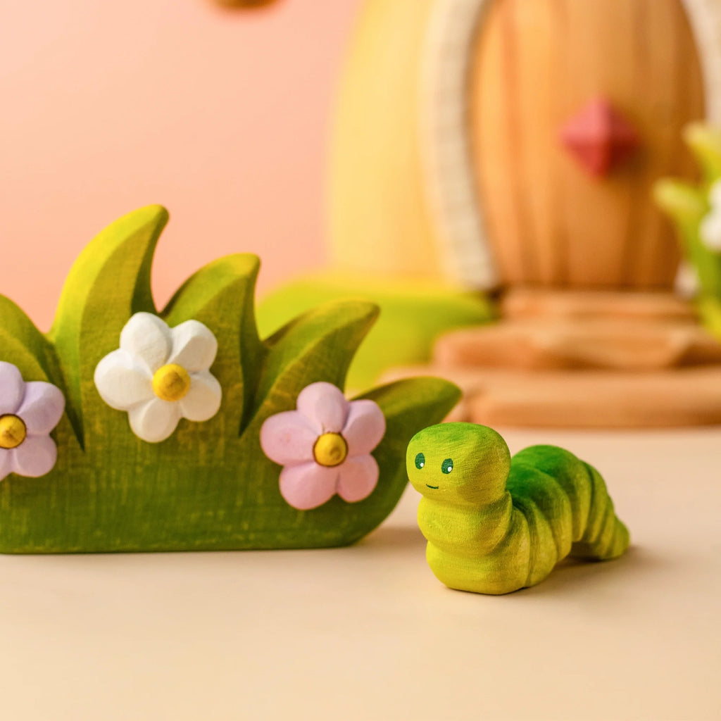 The Bumbu Handmade Wooden Caterpillar, painted in green, rests beside a decorative green piece adorned with white and pink flowers—inviting imaginative play. The softly blurred background features pastel colors and shapes, creating a whimsical scene.