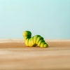 A Bumbu Handmade Wooden Caterpillar, small and green with stripes, sits on a wooden surface against a light blue background.