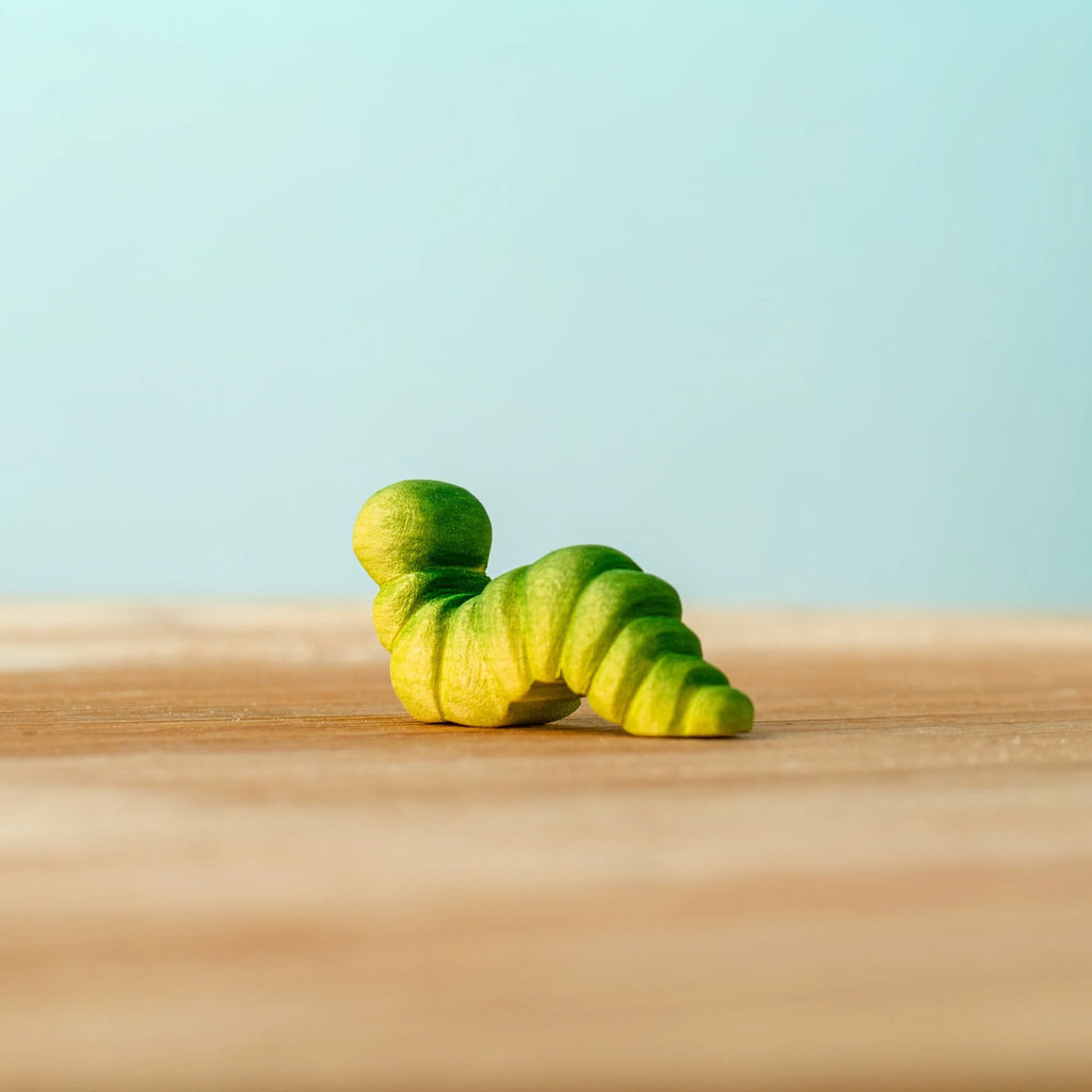 A Bumbu Handmade Wooden Caterpillar, small and green with stripes, sits on a wooden surface against a light blue background.
