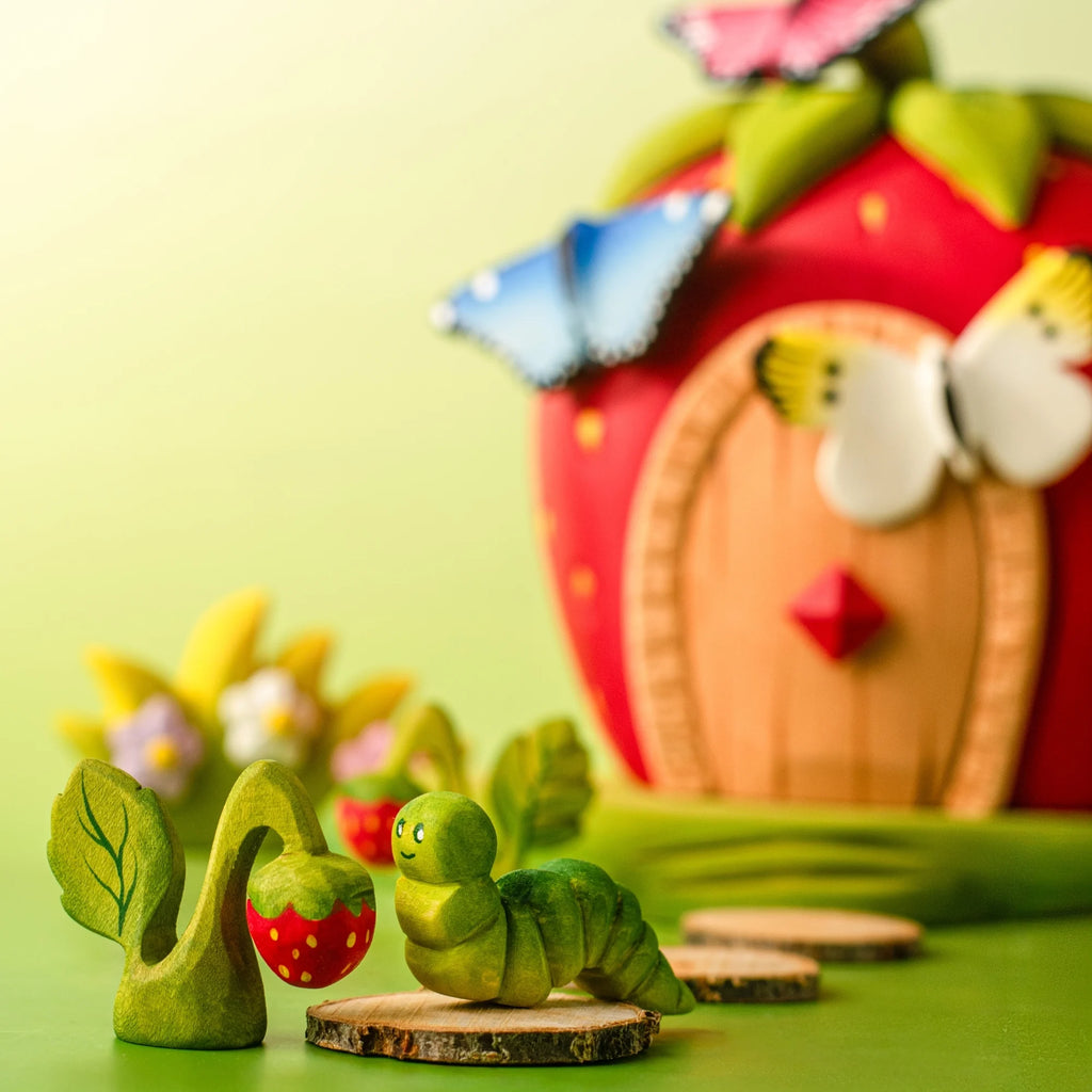 The Bumbu Handmade Wooden Caterpillar, crafted from green wood, sits atop a wooden disk next to a strawberry and leaves, with a charming red strawberry house and butterflies softly blurred in the background—an ideal scene for sparking imaginative play.