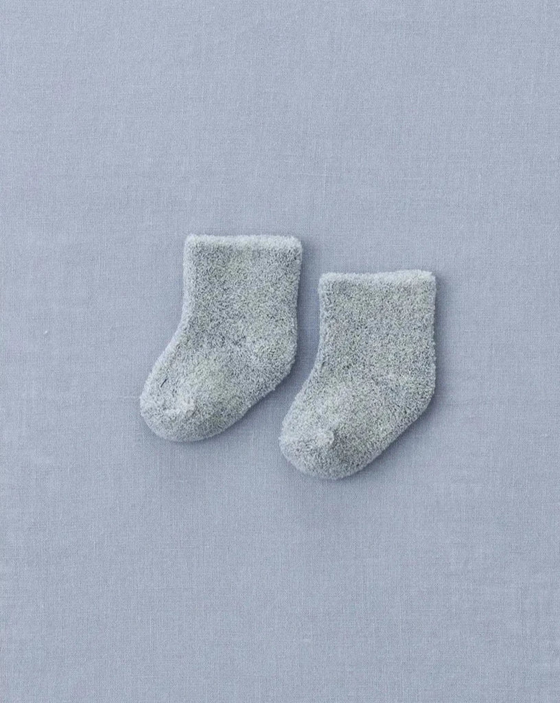 A pair of small, fluffy, gray Baby Pile Knit Socks - Blue is placed on a light blue textured fabric background. The socks are slightly angled with the toes pointing inward.