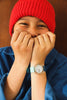 A smiling child in a red knit hat and blue shirt holds their hands to their face, wearing the Mini Kyomo Watch - Blue Ocean with a light blue strap and white dial.