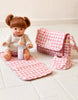 A Tiny Tummies Pink Gift Box - The Essentials by Tiny Harlow features a baby doll with brown hair seated on a pink and white checkered mat, dressed in a snug outfit. Beside the doll is a corresponding checkered diaper bag filled with all the essential items, including a pink bottle positioned nearby. This delightful set is an ideal gift for little ones.