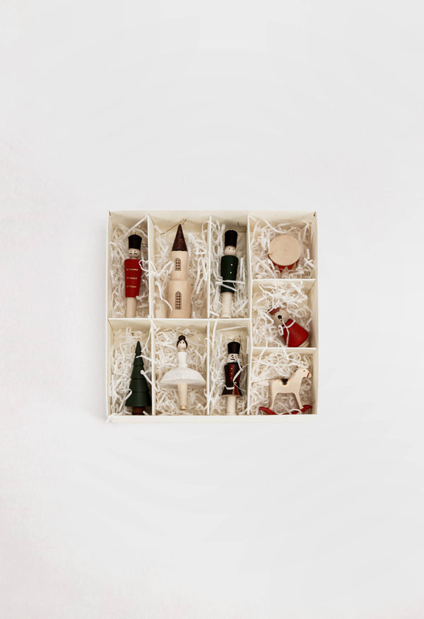 The Sabo Concept Christmas Ornaments set features nine festive hand-painted wooden pieces—like a red bottle, soldier, drum, and star—each showcased in a square box with compartments on white shredded paper against a white background.
