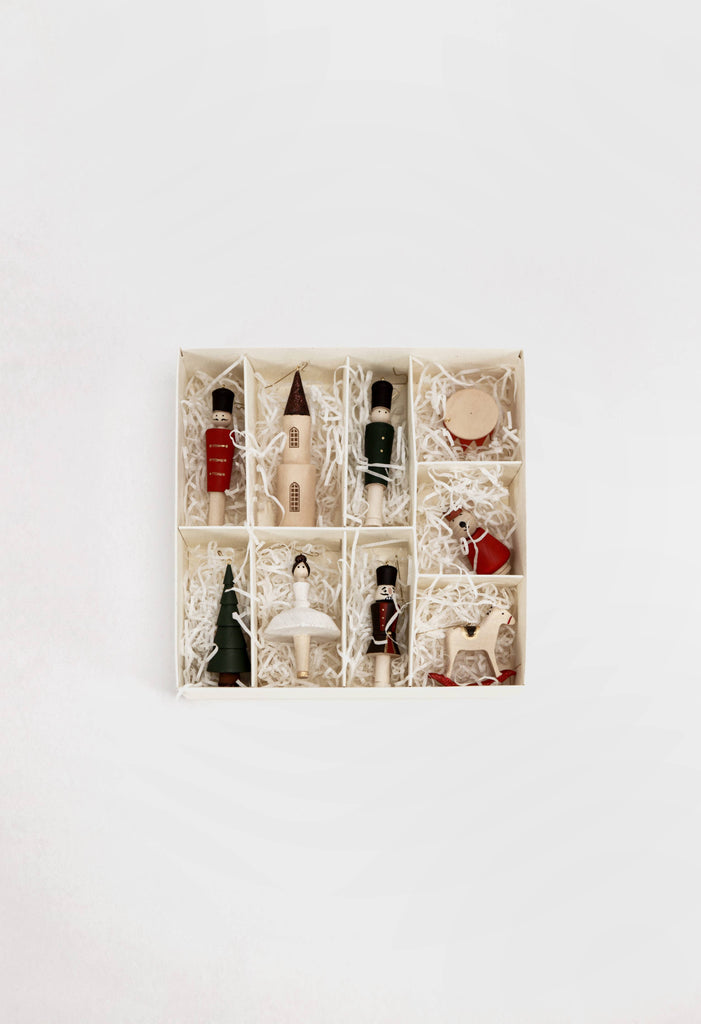 The Sabo Concept Christmas Ornaments set features nine festive hand-painted wooden pieces—like a red bottle, soldier, drum, and star—each showcased in a square box with compartments on white shredded paper against a white background.