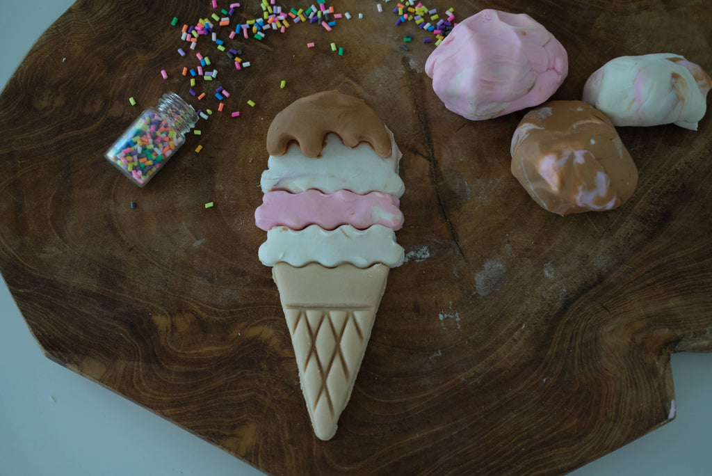 A realistic clay ice cream cone with three scoops rests on a wooden surface, surrounded by colorful sprinkles and marbled clay pieces, inviting sensory play. Perfect for imaginative fun, this scene becomes even more creative with the Ice Cream Cutter Set – Eco Playdough Tool.