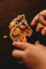 A child’s hands press a cow-shaped cutter from the Farm Cutter Set - Eco Playdough Tool into orange and yellow dough on a dark wooden surface—ideal for sustainable sensory play.