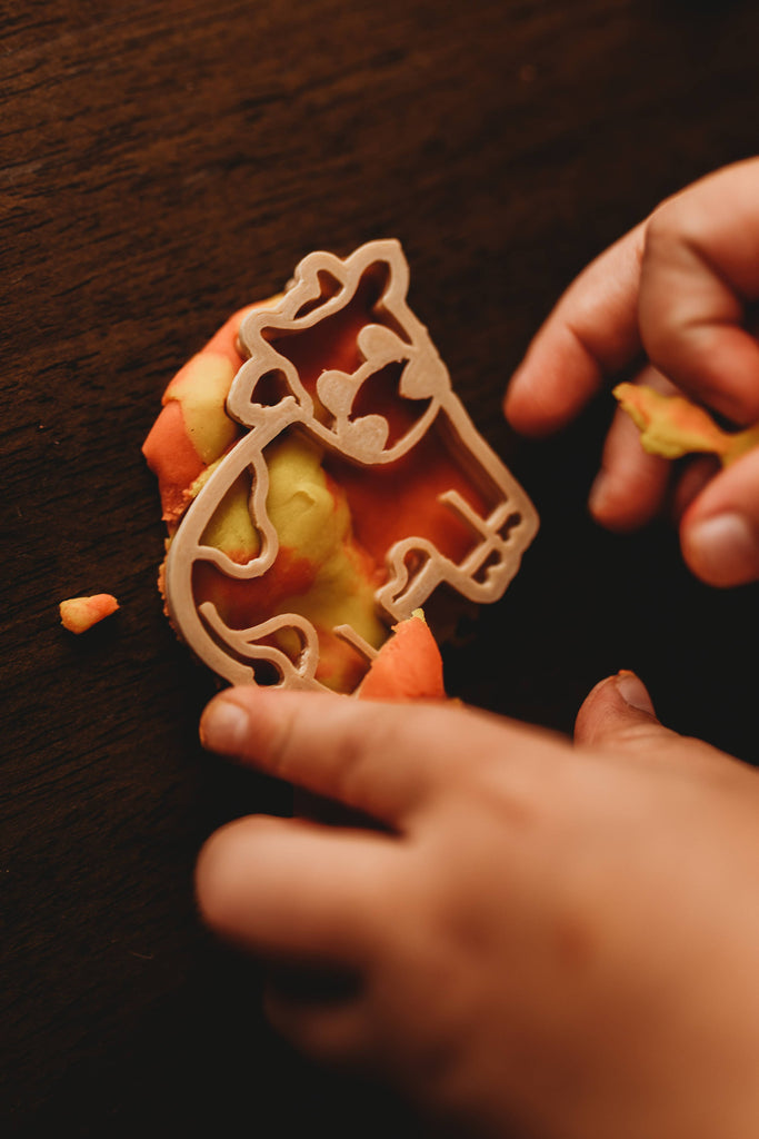 A child’s hands press a cow-shaped cutter from the Farm Cutter Set - Eco Playdough Tool into orange and yellow dough on a dark wooden surface—ideal for sustainable sensory play.