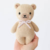 A hand holds the Cuddle + Kind Baby Honey Bear, an heirloom-quality hand-knit teddy bear with black eyes, a black nose, a smiling mouth, and a lavender bow around its neck, shown against a plain white background.