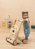 A smiling toddler in blue overalls stands on a wooden floor, holding the Wooden Activity Walker. A baby walker and a wooden activity cube toy, designed as engaging sensory toys, are visible in the background.