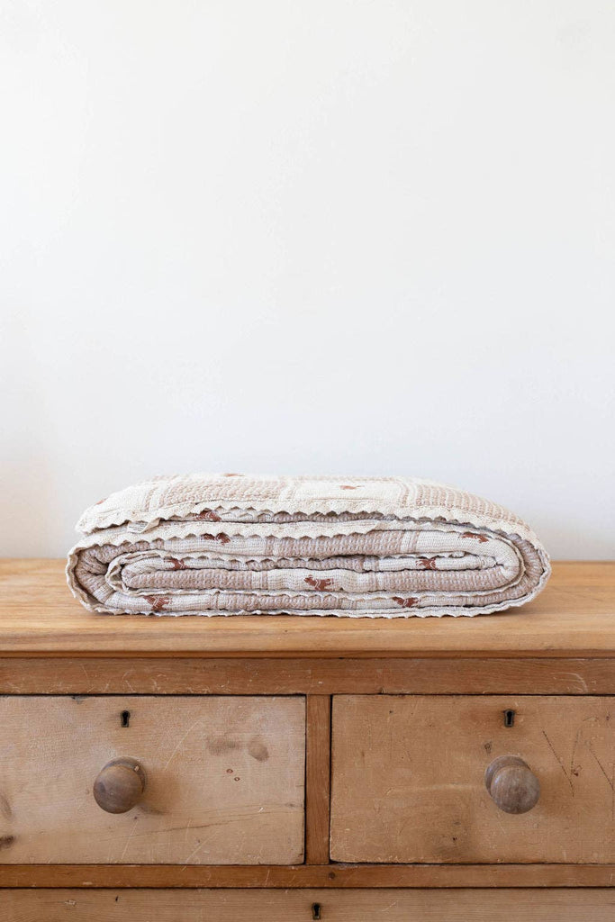 The Organic Cotton Twin Size Blanket - Pony, cream-colored with subtle patterns, is neatly folded atop a wooden dresser with two drawers and round knobs, set against a plain light wall.