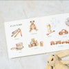 Our Big Book of First Words is open on a marble surface, displaying watercolor illustrations labeled with words like crayons, teddy bear, and toy car. A plush bunny nearby makes it perfect for building vocabulary with little ones.