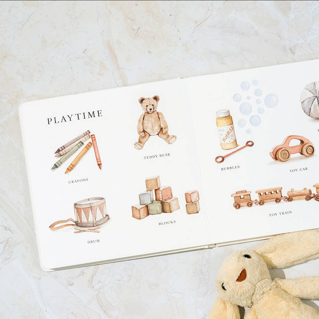 Our Big Book of First Words is open on a marble surface, displaying watercolor illustrations labeled with words like crayons, teddy bear, and toy car. A plush bunny nearby makes it perfect for building vocabulary with little ones.