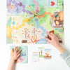 An overhead view of a colorful cooperative children's board game with a forest theme spread out on a table, featuring the Slumberkins Feelings Adventure Board Game designed to enhance emotional regulation skills. A child's hand touches