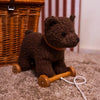 A plush brown Pull-Along Raoul bear toy with wheels and a pull string sits on a carpeted floor next to a wicker basket. This charming Pull-Along Raoul encourages the development of gross motor skills as children engage in active play. Vertical striped wallpaper is in the background.