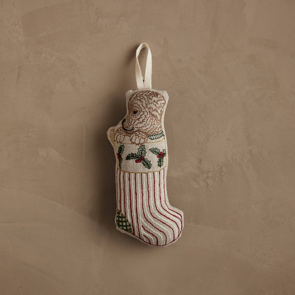 The Coral & Tusk Puppy Stocking Ornament, featuring red stripes, a green patch, holly embroidery, and a dog in a sweater, hangs by a white loop—a charming holiday decoration for your home.