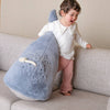 A smiling young child stands on a beige couch, leaning against the Ride-On Whale Pouf—a plush blue pouf featuring a playful whale design and rope handle—radiating happiness and playful energy.