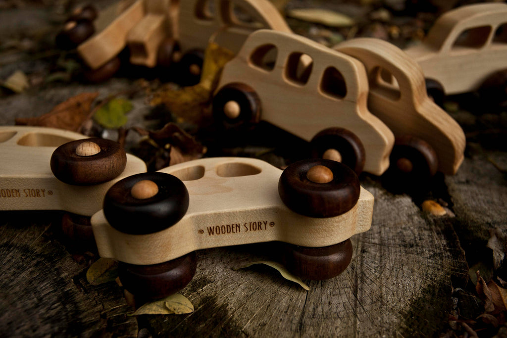 Several Little Wooden French Cars, each handcrafted from natural wood with dark wheels and a smooth finish, rest outdoors on a rustic surface among dry leaves. Each eco-friendly baby toy is branded "WOODEN STORY.