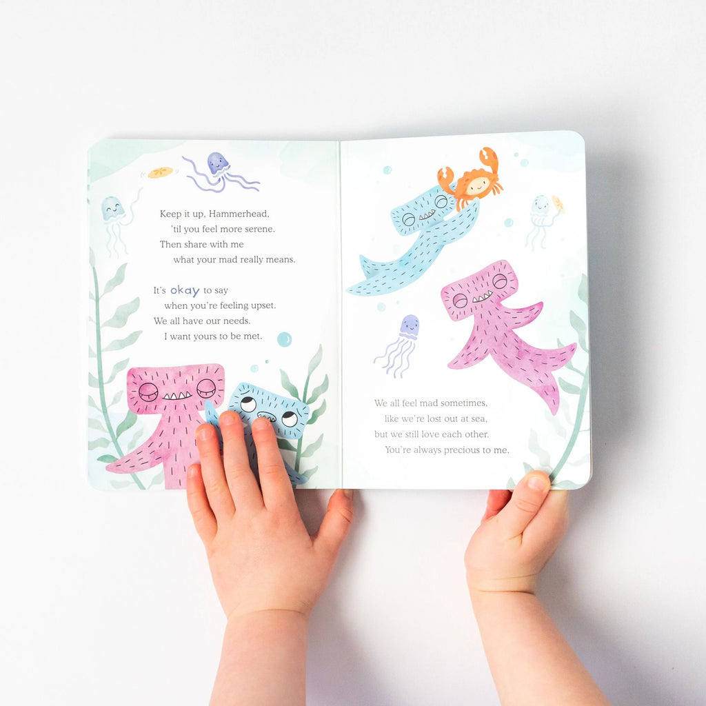 A child holds open a colorful children's book featuring playful sea creatures, including two pink octopuses, a blue whale, a blue hammerhead shark, and a crab. The text on the pages provides reassuring and comforting messages to help with emotional regulation and conflict resolution. The book is part of the Slumberkins Hammerhead Kin + Lesson Book - Conflict Resolution set.