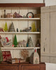 A wooden cabinet with open doors shows Christmas decor—mini trees, gifts, birds, a sleigh, a lamb, and the Coral & Tusk Home for the Holidays Tissue Box Cover shaped like a house—all arranged on white shelves.