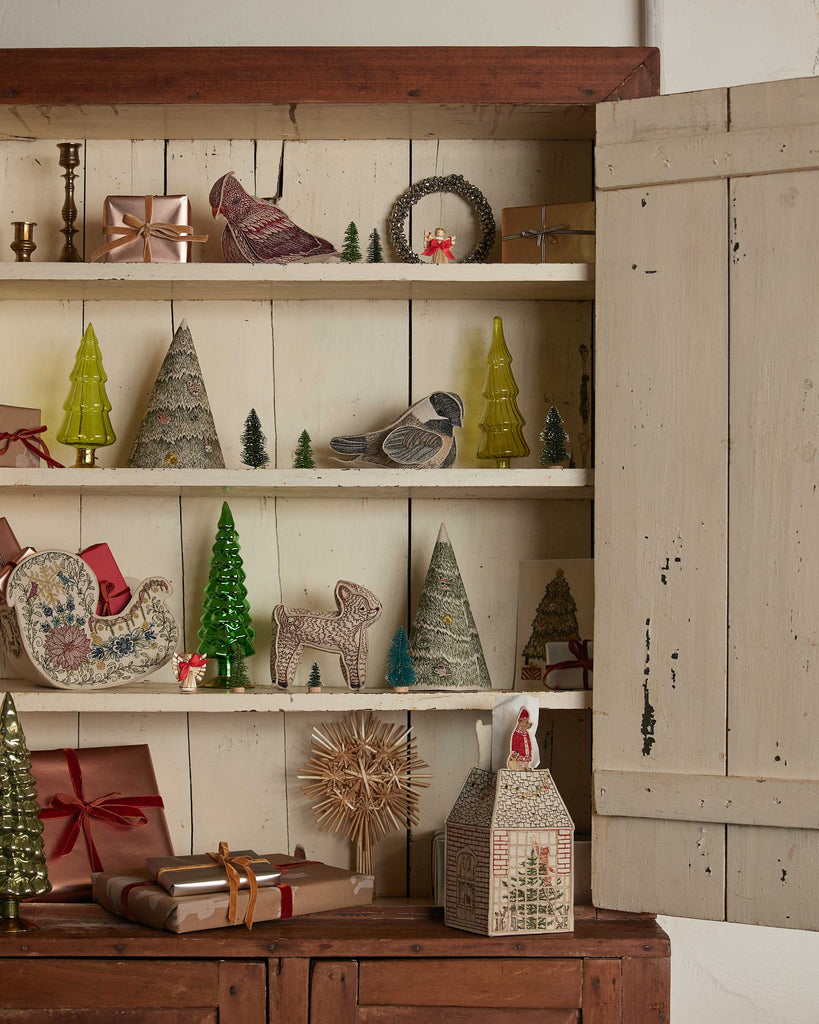 A wooden cabinet with open doors shows Christmas decor—mini trees, gifts, birds, a sleigh, a lamb, and the Coral & Tusk Home for the Holidays Tissue Box Cover shaped like a house—all arranged on white shelves.