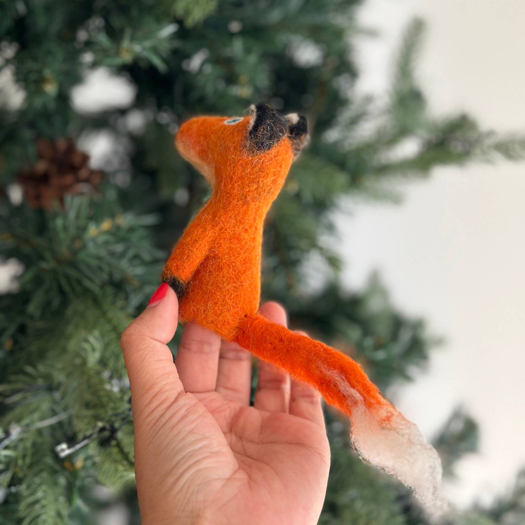 A hand holds the enchanting Fox Finger Puppet, crafted from wool felt and shaped like an orange fox with a long tail, set against the backdrop of a green pine tree, perfect for enhancing storytelling moments. Note: This item ships in approximately one week.