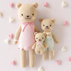 The Cuddle + Kind Baby Honey Bear (Lavender Bow), a hand-knit, heirloom-quality doll, is displayed on a light background with pastel heart decorations for a charming nursery accent.