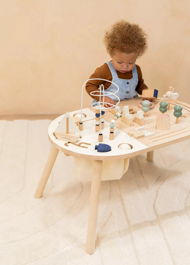 A toddler with curly hair plays intently with a bead maze and various wooden toys on a Wooden Activity Table in a softly lit room with a beige background.