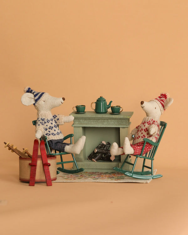 The Maileg Ski Lounge Set depicts two plush mice dressed in charming winter outfits, relaxing in rocking chairs by a tiny fireplace. They are holding cups, with a teapot placed on the mantle and a basket of wrapped items close by. It's important to be mindful of small parts that could pose a choking hazard against the warm beige backdrop.