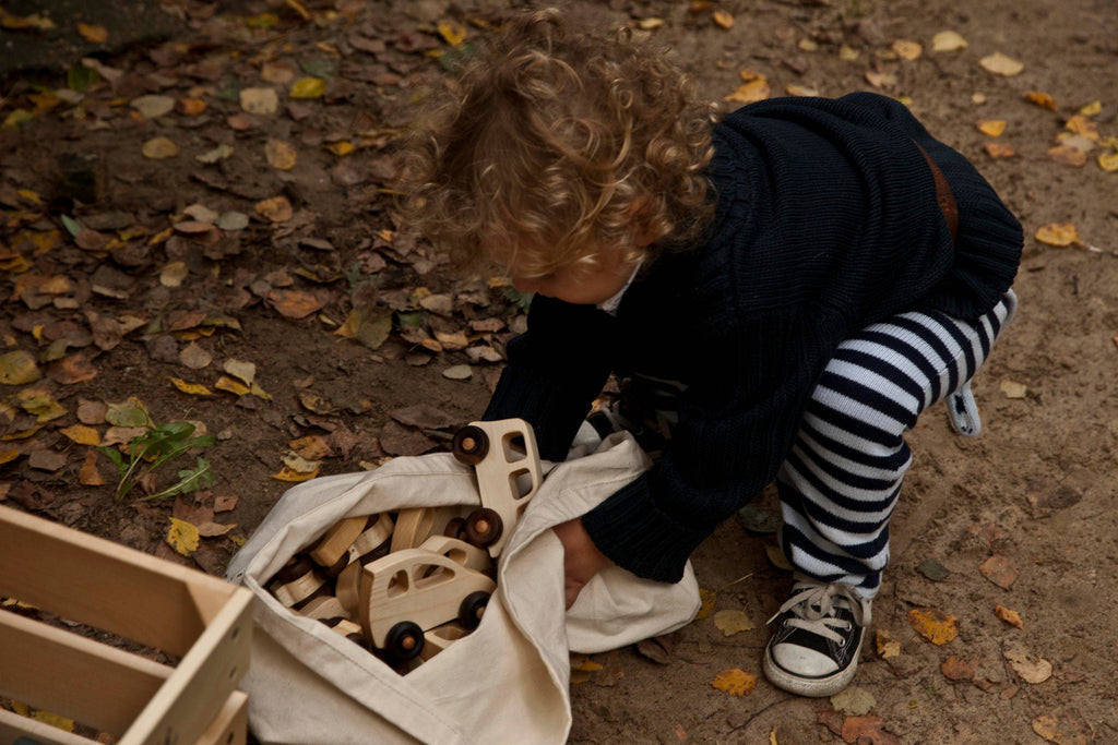 A young child with curly hair, in a dark sweater and striped pants, crouches outdoors on fallen leaves, taking the Little Wooden French Car from a cloth bag.